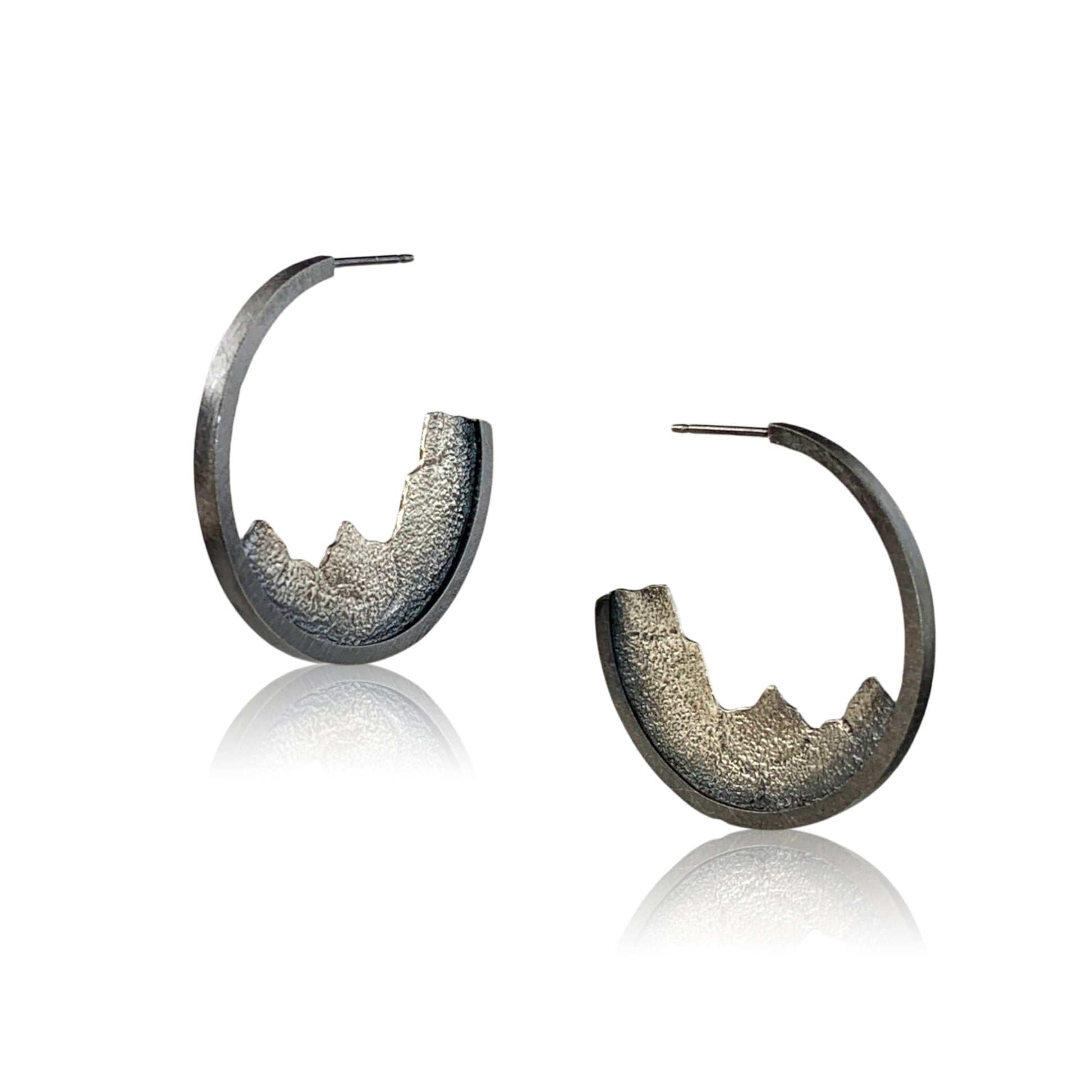 A front/vertical view of two open-hoop earrings with silver posts. Nestled into the bottom halves are textured ombre silver pieces, like slices of a mountain range.