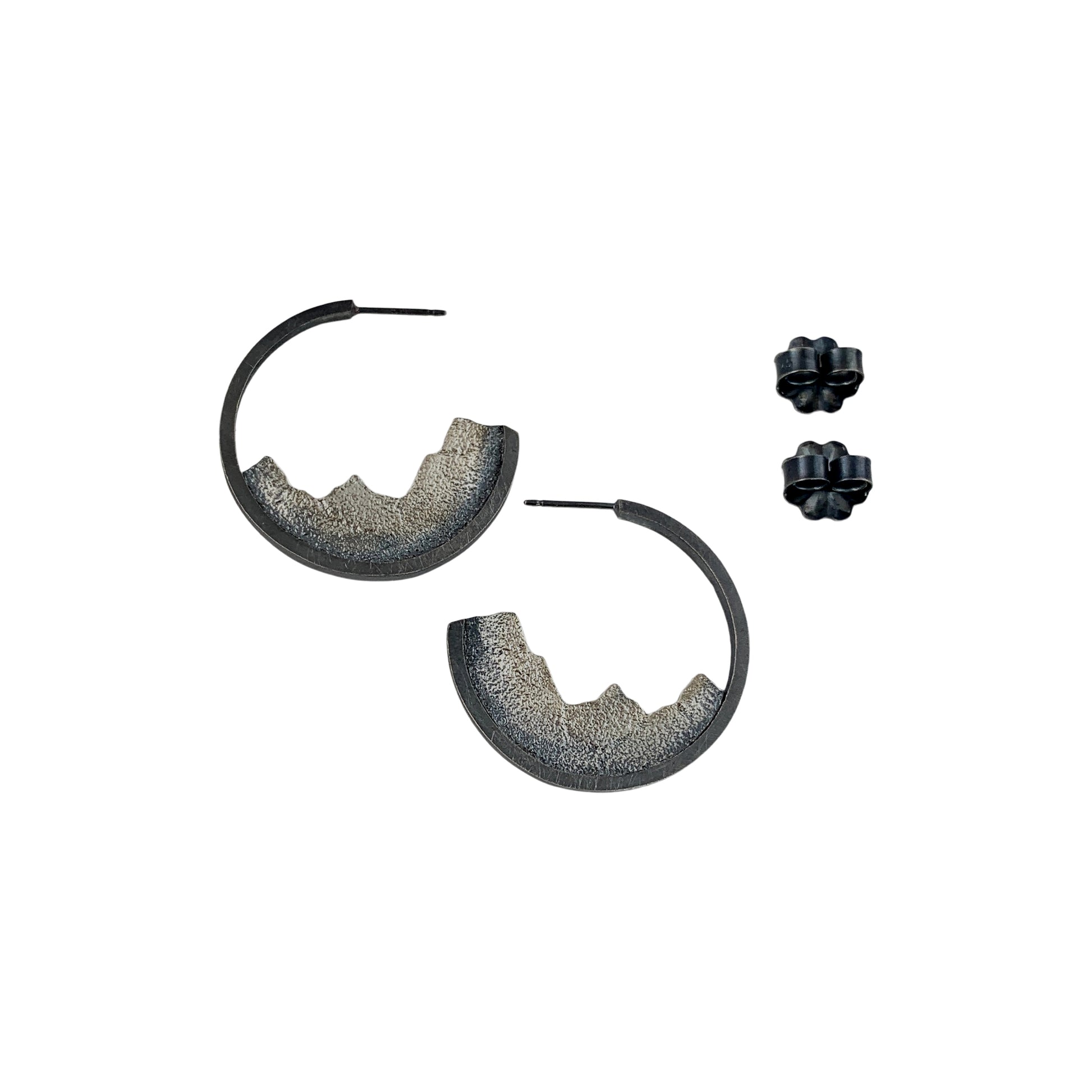 A vertical view of two open-hoop earrings with silver posts. Nestled into the bottom halves are textured ombre silver pieces, like slices of a mountain range. Two fasteners are shown to the side.