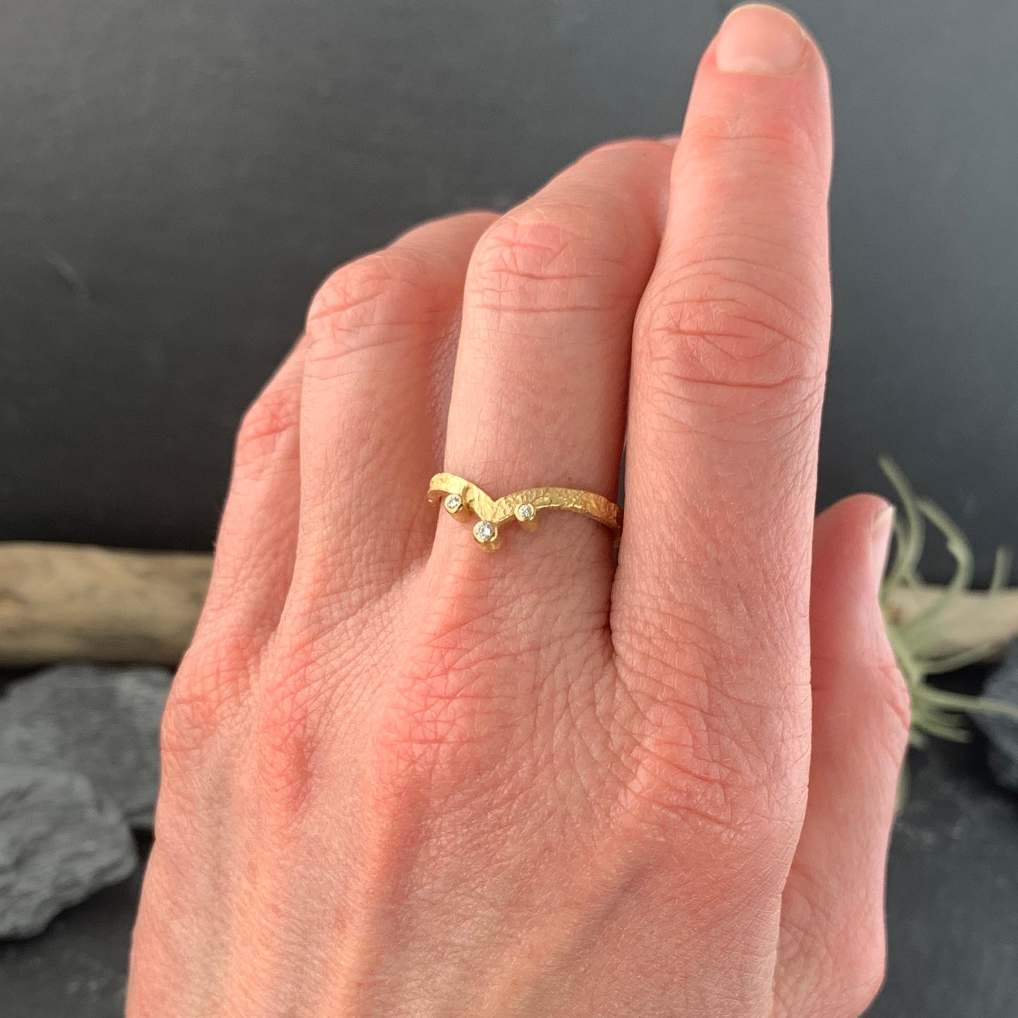 View of a woman's left hand with the ring on her middle finger. The ring is a pounded texture gold ring that dips down to a V shape. Three gold-wrapped white diamonds accent the bottom and sides of the V.