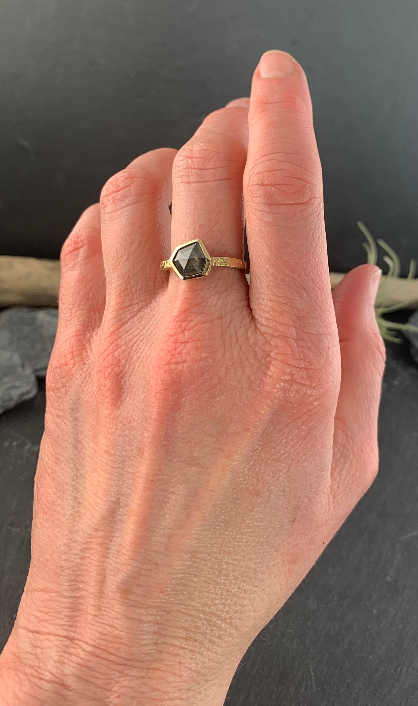 A woman's left hand, wearing the ring on her middle finger. The ring is a narrow, textured gold band. At the center is a gray speckled six-irregular-sided diamond.The diamond is encased in a smooth gold wrapping.