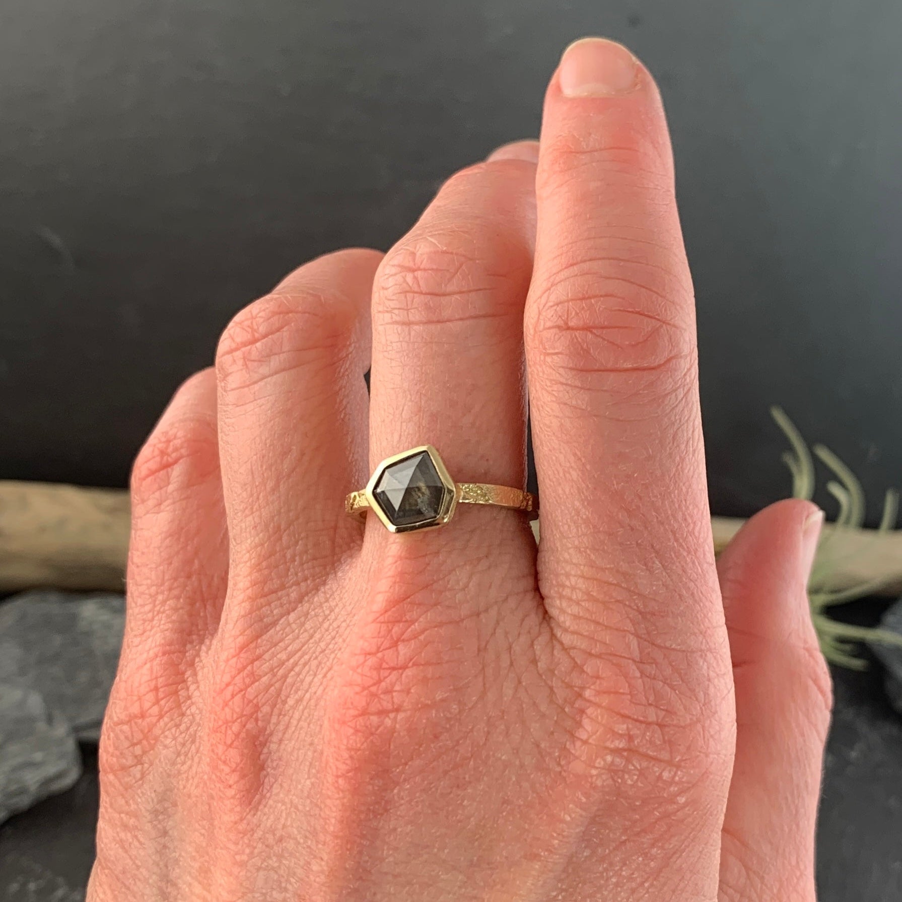 A woman's left hand, wearing the ring on her middle finger. The ring is a narrow, textured gold band. At the center is a gray speckled six-irregular-sided diamond.The diamond is encased in a smooth gold wrapping.
