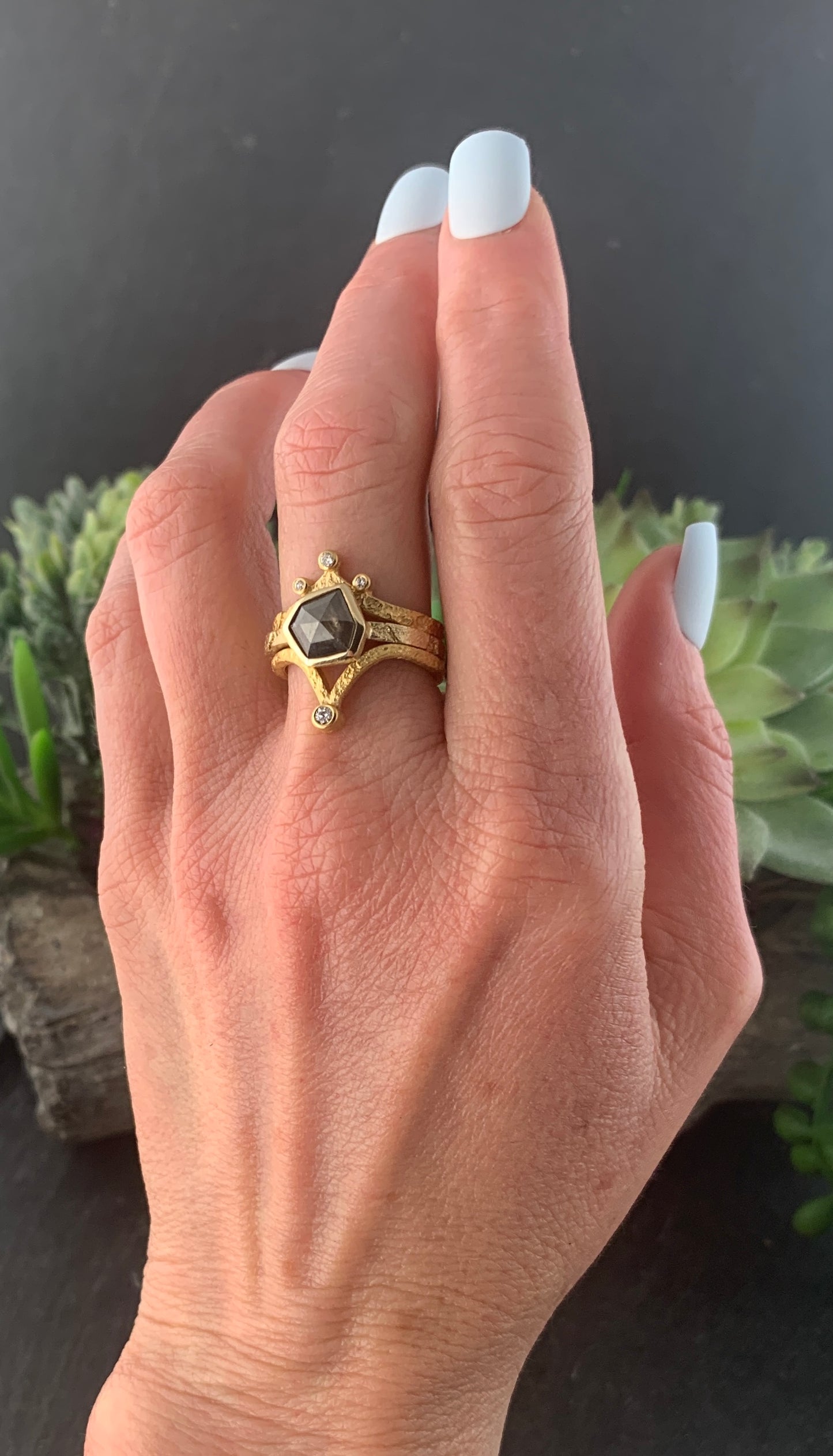A woman's left hand, wearing the Bedrock ring on her middle finger. The ring is a narrow, textured gold band. At the center is a gray speckled six-irregular-sided diamond.The diamond is encased in a smooth gold wrapping. Stacked on each side of the ring are two other gold and diamond rings, demonstrating the Bedrock ring's versatility.