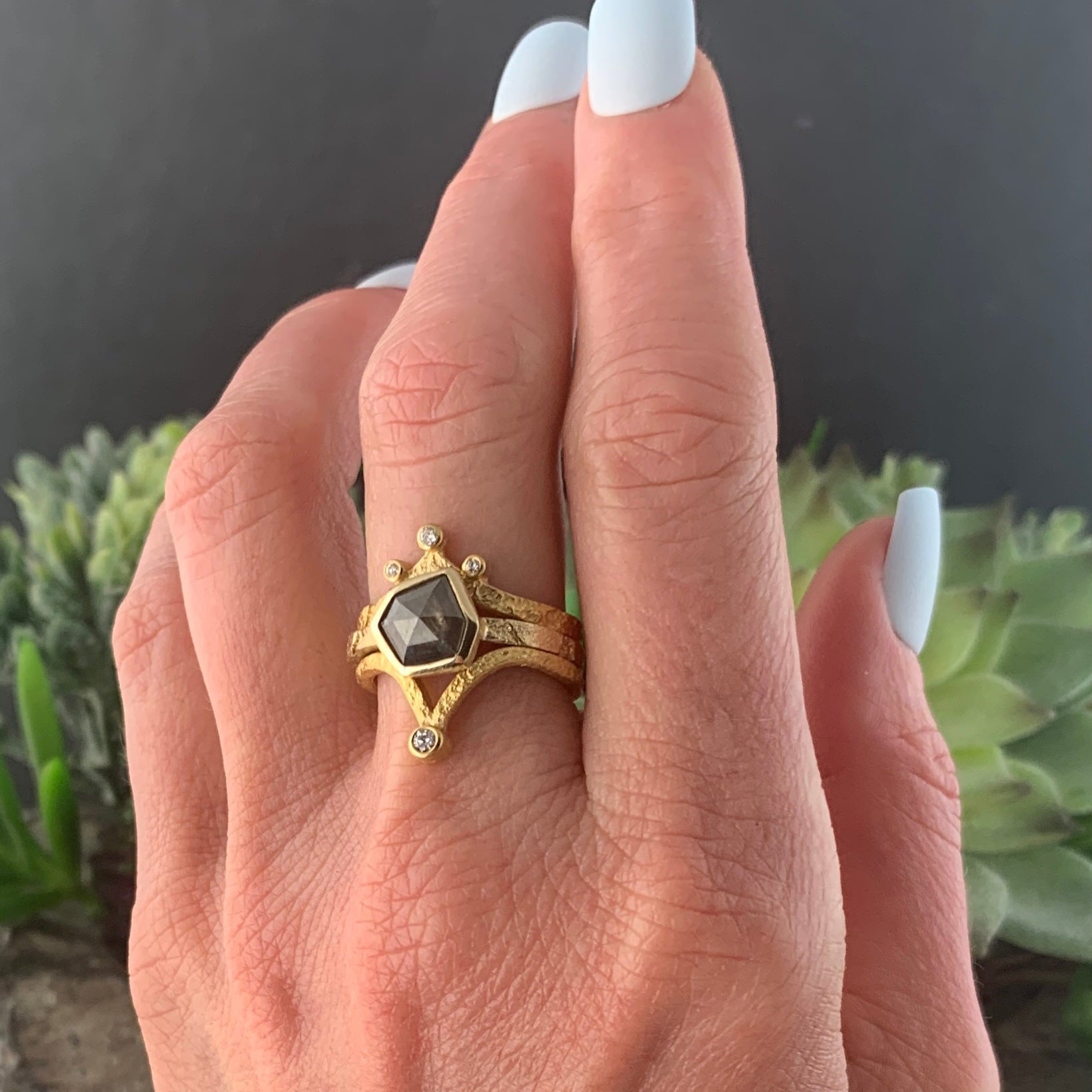 A woman's left hand, wearing the Bedrock ring on her middle finger. The ring is a narrow, textured gold band. At the center is a gray speckled six-irregular-sided diamond.The diamond is encased in a smooth gold wrapping. Stacked on each side of the ring are two other gold and diamond rings, demonstrating the Bedrock ring's versatility.