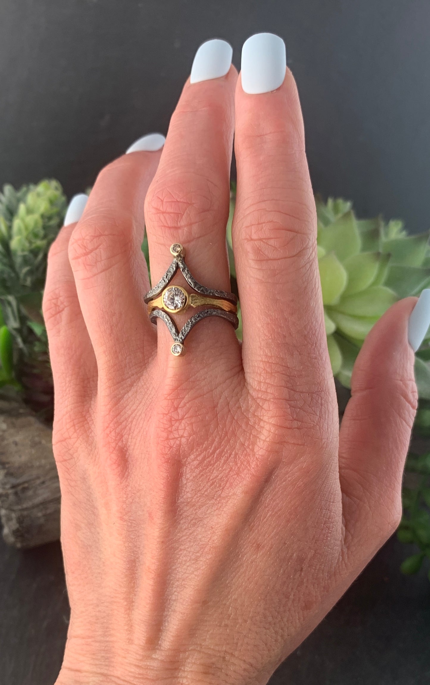 A woman's left hand, wearing two textured dark gray bands with V shapes. At the bottom of each V is a yellowish diamond, set in smooth yellow gold. Between these two rings is a gold band with a large white diamond, demonstrating the stackability of this ring.