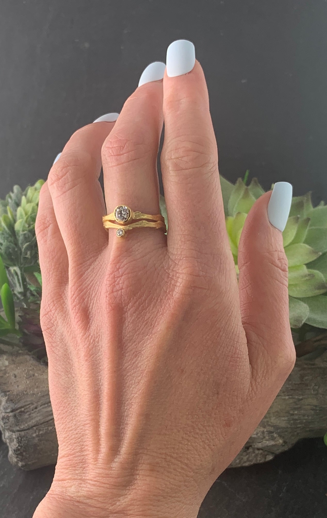 A woman's left hand, wearing a gold ring with a wavy, textured band. On the lower edge of the band is a diamond set in gold. Stacked above this ring is another gold ring with a bigger diamond, demonstrating the Cypress ring's versatility.